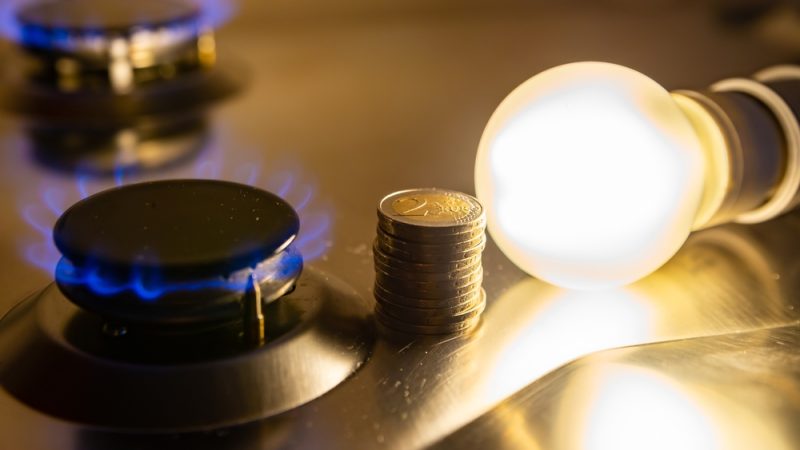 Der Zusammenhang der vom Gaspreis in Österreich und dem Strompreis. Eine eingeschaltete Gas-Herdplatte, ein Stapel 2 Euro Münzen und eine eingeschaltete Glühbirne.